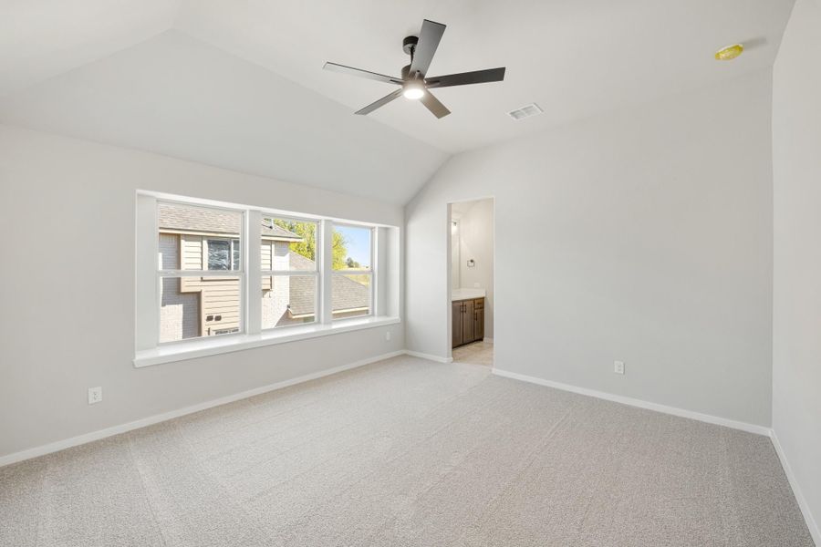 Empty master bedroom with vaulted ceiling, ceiling fan, carpet flooring, and ensuite bathroom doorway