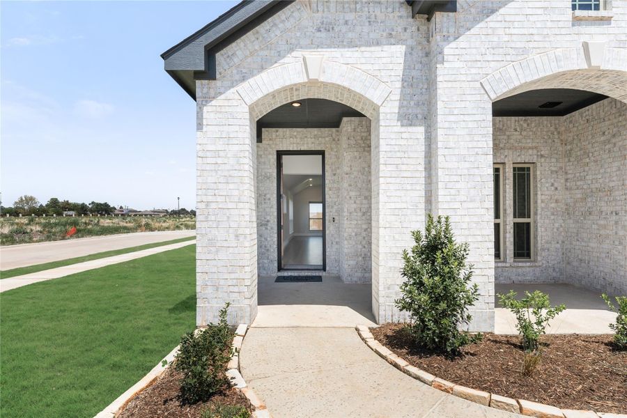 Home entrance with white brick arched portico, modern front door, concrete walkway, and landscaped shrubs