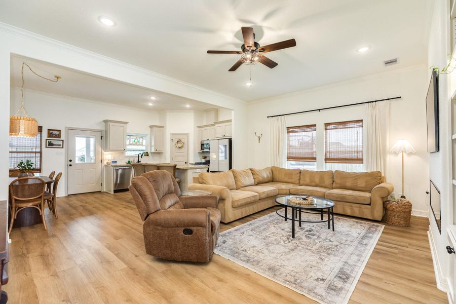 This photo showcases a spacious open-concept living area with warm wood flooring and modern ceiling fan. The adjacent kitchen has light cabinetry, stainless steel appliances, and a breakfast bar. Natural light fills the space through large windows with bamboo shades.