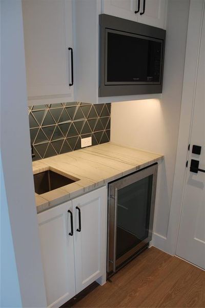 Kitchen with light stone countertops, wine cooler, backsplash, wood-type flooring, and white cabinets