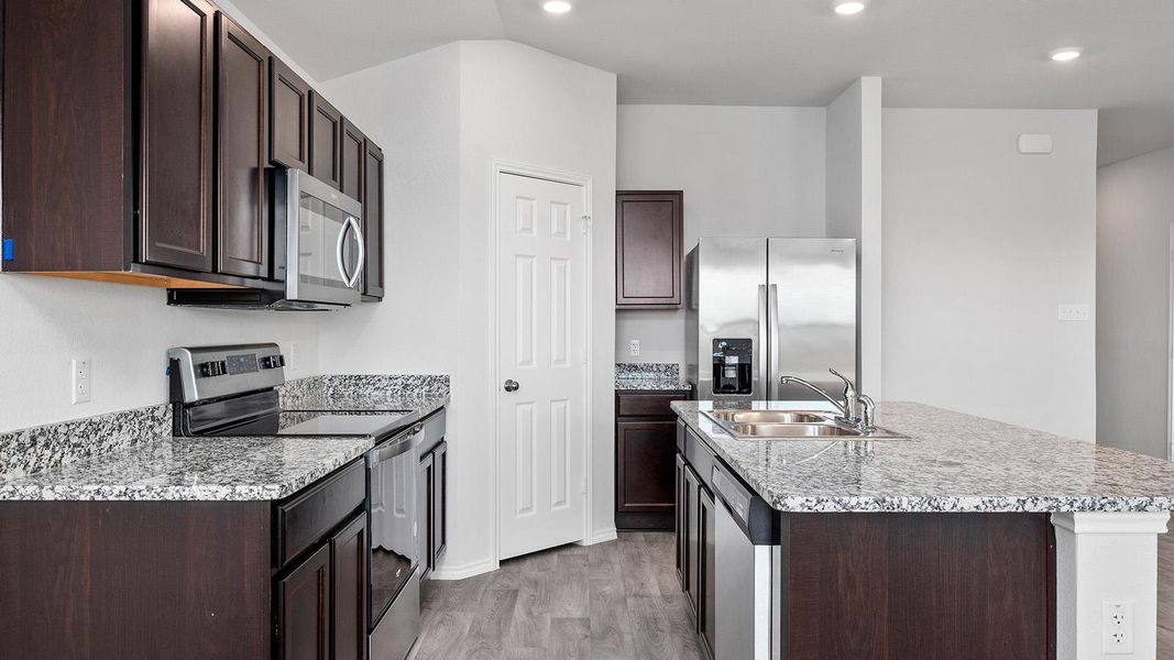 Kitchen with appliances with stainless steel finishes, dark brown cabinets, an island with sink, light wood-style flooring, and light stone countertops
