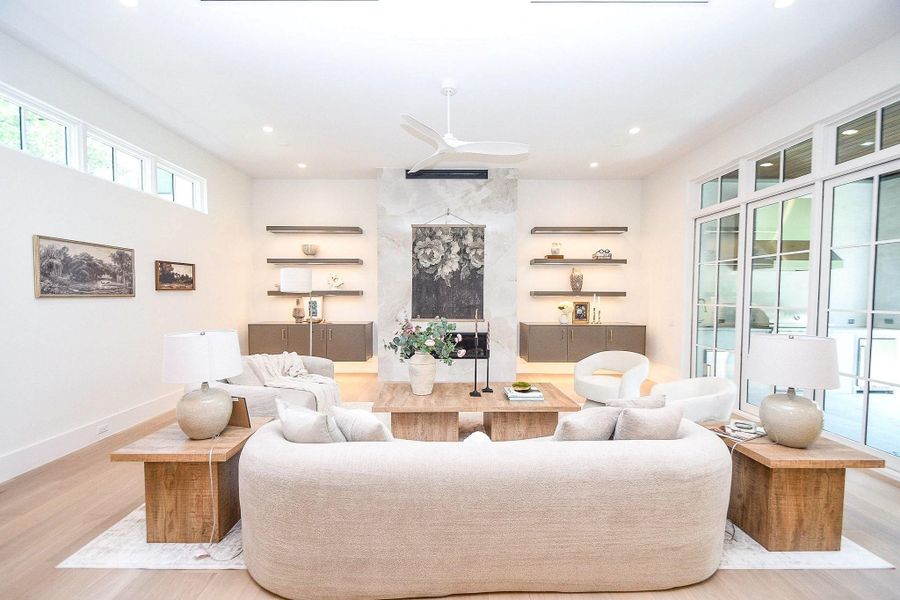 This spacious living room features a modern design with high ceilings, large windows, and abundant natural light. It has a cozy seating arrangement with neutral-toned furniture, a central coffee table, built-in shelving, and elegant decor. The room offers a seamless indoor-outdoor flow through large glass doors.