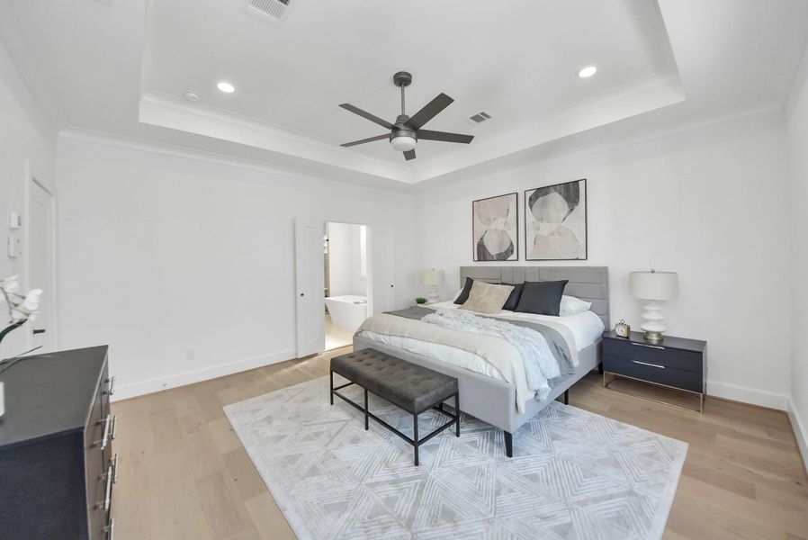 This spacious, modern bedroom invites relaxation with a neutral color palette, tray ceiling, recessed lighting, and a ceiling fan. A large bed with stylish bedding and matching nightstands anchors the room, while a contemporary rug adds warmth. The room connects seamlessly to an en-suite bathroom, enhancing the suite’s luxurious feel.