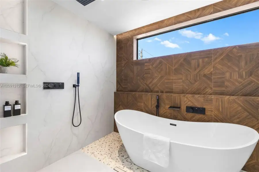 Freestanding soaking tub featuring matte black wall-mounted fixtures, geometric wood-patterned accent wall, recessed shelving, and a panoramic clerestory window Freestanding soaking tub featuring matte black wall-mounted fixtures, geometric wood-patterned accent wall, recessed shelving, and a panoramic clerestory window