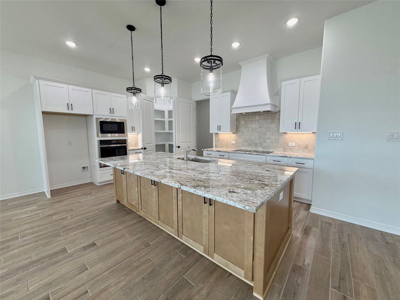 Spacious kitchen featuring an expansive center island with stone countertop, wood-finish flooring, custom cabinetry, built-in stainless steel appliances, and a tile backsplash