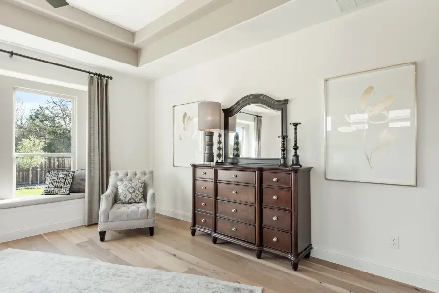 Bedroom with dark wood dresser and mirror, tufted accent chair, window seat, and light hardwood floors