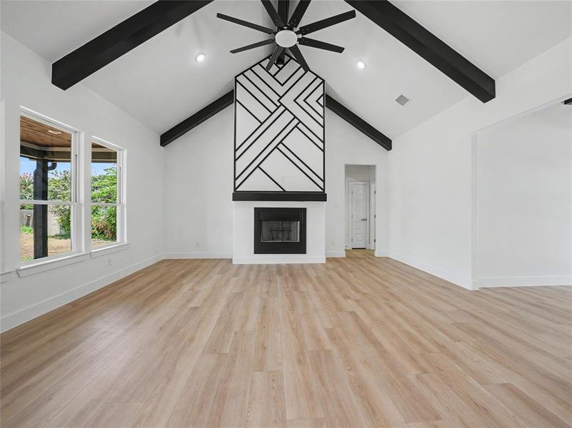 Spacious living area featuring a vaulted ceiling with exposed black beams, a decorative fireplace with a geometric mantel, and wood-finish flooring Spacious living area featuring a vaulted ceiling with exposed black beams, a decorative fireplace with a geometric mantel, and wood-finish flooring