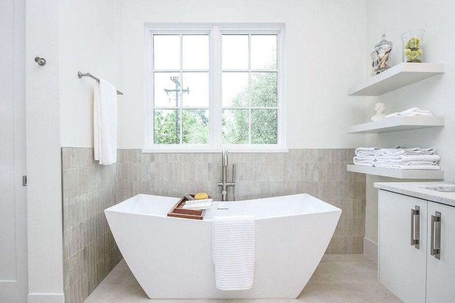 This bathroom features a modern, freestanding bathtub with a sleek design, set against neutral tile and white walls. A large window offers natural light, while open shelving provides stylish storage for towels and decor.