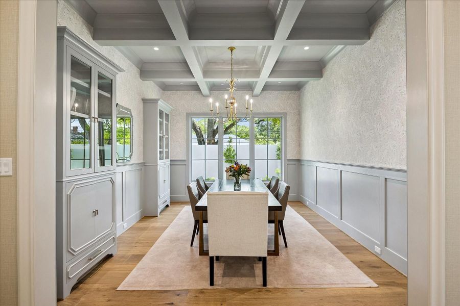 Formal dining room featuring a striking coffered ceiling, classic candelabra-style chandelier, gorgeous wallpaper and wainscotting. Fantastic custom built-in cabinetry and large windows create a bright, sophisticated setting for dining and entertaining.