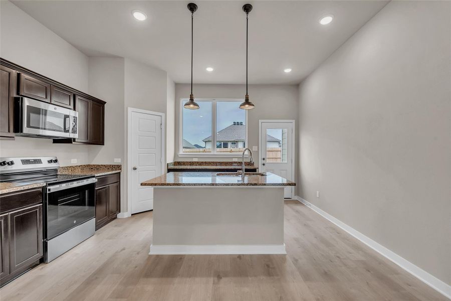 Kitchen featuring stainless steel appliances, dark stone countertops, dark wood finish cabinetry, decorative light fixtures, and light wood finished floors