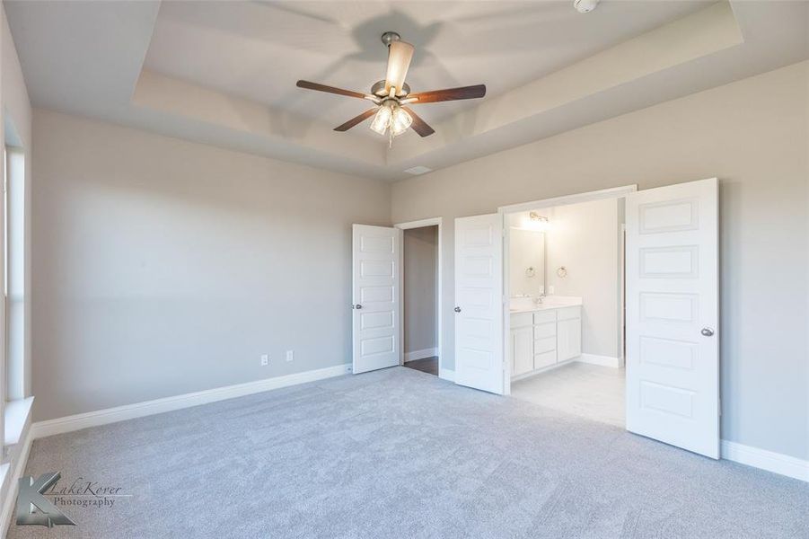 Unfurnished bedroom with a raised ceiling, light colored carpet, ceiling fan, and connected bathroom