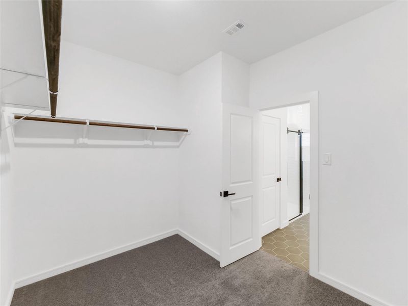 A spacious walk-in closet with a single wooden shelf and hanging bar. It has clean white walls, carpeted flooring, and a door leading to a bathroom with hexagonal tile flooring.