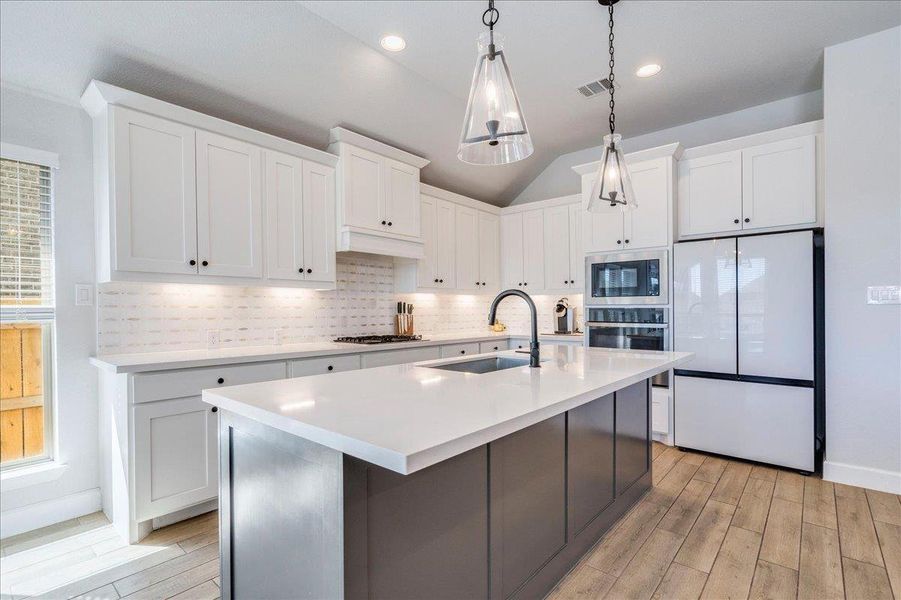 Dual tone kitchen featuring stainless steel appliances, a center island with sink, hanging light fixtures, light wood finished floors, and lofted ceiling