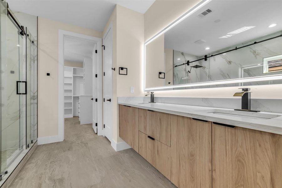 This modern primary bathroom features a spacious double vanity with sleek wood cabinetry and a large, illuminated mirror. It has a glass-enclosed shower and ample storage space, providing a clean and contemporary look.