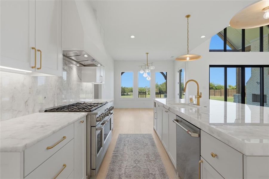 Gourmet kitchen featuring a stainless steel gas range, large island with integrated sink, white cabinetry with gold-tone hardware, full-height stone slab backsplash, and wood-finish flooring