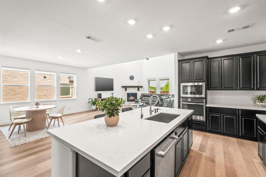 Kitchen featuring open floor plan, dark cabinetry, light wood-style floors, stainless steel appliances, and a kitchen island with sink