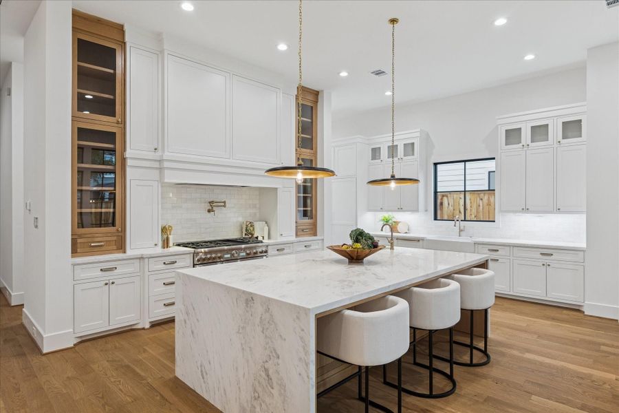 Floor-to-ceiling millwork frames a professional-grade Thermador range beneath a sculpted hood, flanked by vertical glass cabinets for elegant display. Brass pendants cast a warm glow across the marble surfaces, while the Pella window package and open layout ensure a bright, functional cooking space. *Previous Project*
