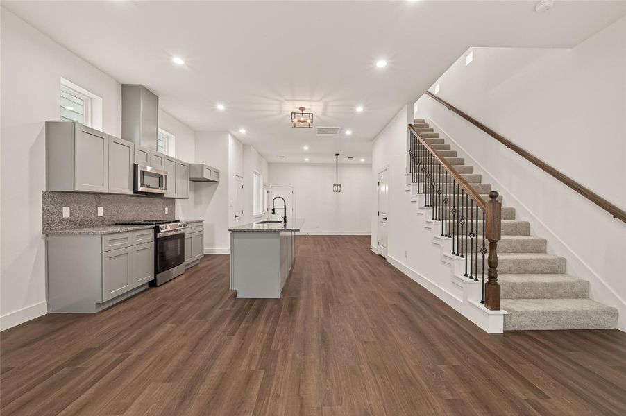 This photo showcases a modern, open-concept kitchen and living area with sleek gray cabinetry, stainless steel appliances, and a large island. The space features warm wood flooring and a carpeted staircase with elegant railing, leading to the upper level. Bright and inviting, it offers a spacious setting for family gatherings and entertaining. This photo showcases a modern, open-concept kitchen and living area with sleek gray cabinetry, stainless steel appliances, and a large island. The space features warm wood flooring and a carpeted staircase with elegant railing, leading to the upper level. Bright and inviting, it offers a spacious setting for family gatherings and entertaining.