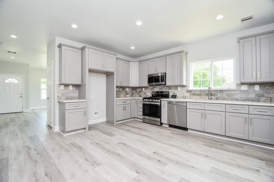 The photo showcases a spacious, modern kitchen with light gray shaker cabinetry, stainless steel appliances, a subway tile backsplash, and sleek, light wood-look flooring. The room is well-lit with recessed lighting and natural light from windows.