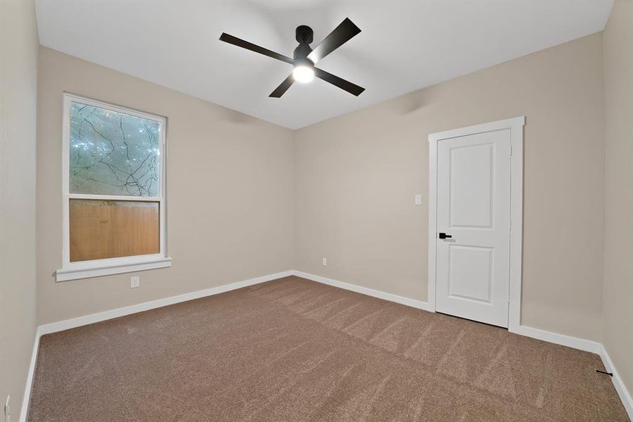 Spacious room featuring neutral paint, brown carpeting, a modern ceiling fan with lighting, a window with white trim, and a white two-panel door with black hardware Spacious room featuring neutral paint, brown carpeting, a modern ceiling fan with lighting, a window with white trim, and a white two-panel door with black hardware