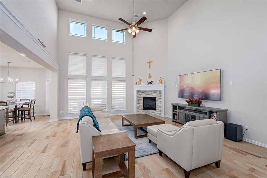 Living room with a stone fireplace, ceiling fan with notable chandelier, a high ceiling, and light wood-type flooring