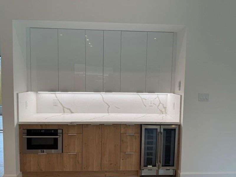 Sleek kitchen area featuring modern cabinetry, a built-in microwave, dual wine fridges, and elegant Quartz backsplash with under-cabinet lighting.