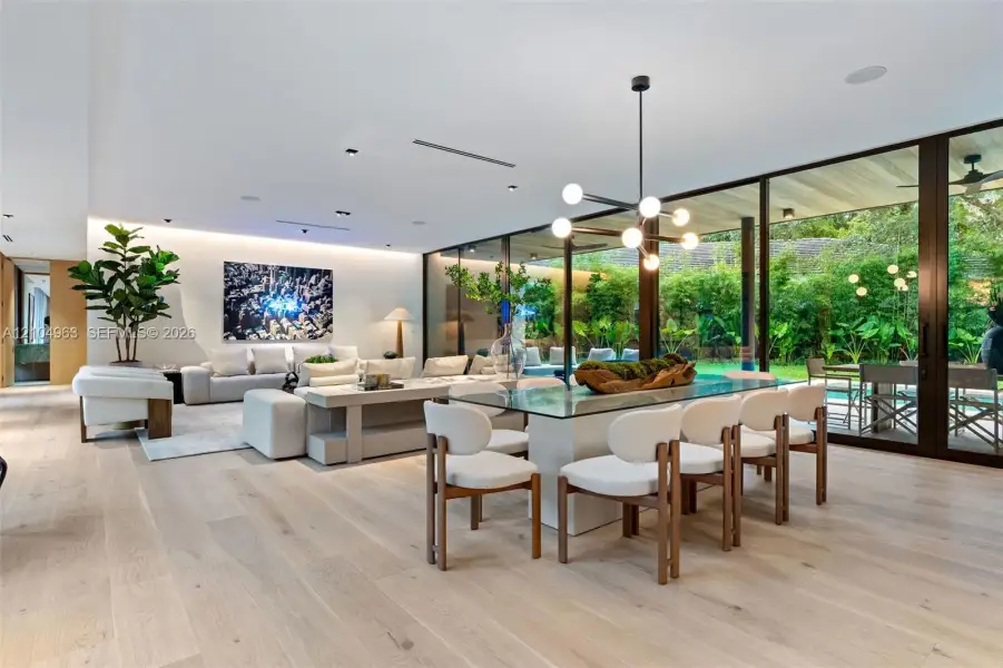 Open-concept interior featuring light wood-finish flooring, floor-to-ceiling glass panel walls, recessed architectural lighting, and a modern multi-arm chandelier