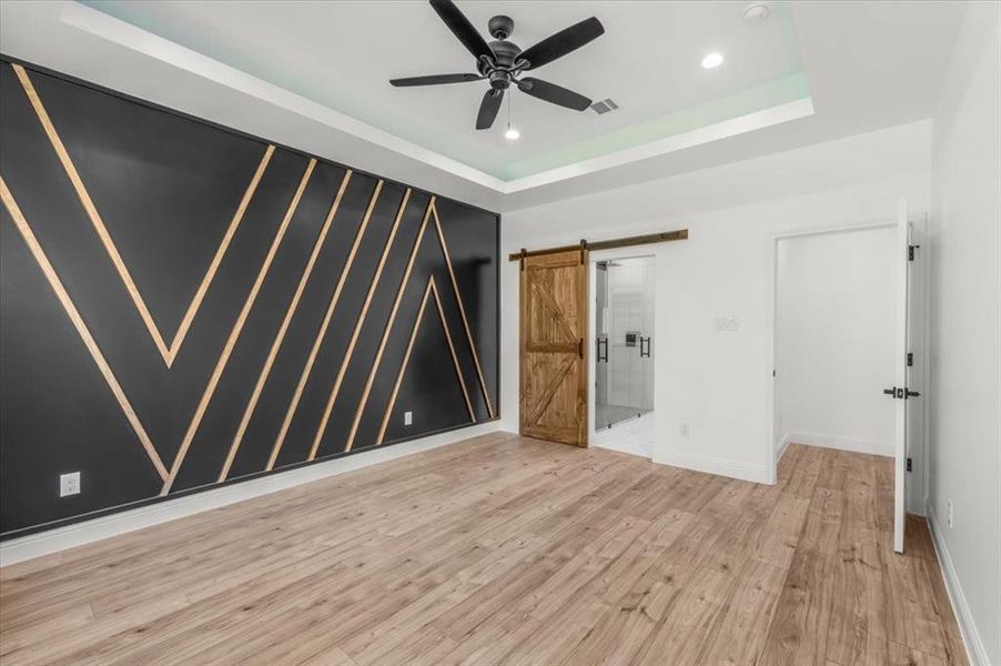 Unfurnished bedroom with a tray ceiling, wood finished floors, a barn door, ensuite bath, and ceiling fan