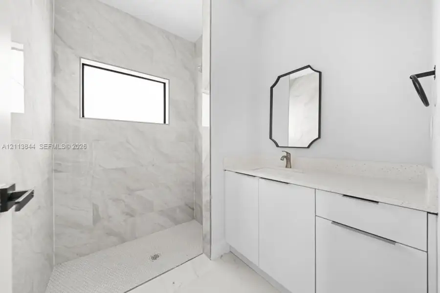 Walk-in shower featuring large-format stone-tile surround, light-colored quartz vanity countertop, white flat-panel cabinetry, hexagonal floor tiling, and dark-finish hardware Walk-in shower featuring large-format stone-tile surround, light-colored quartz vanity countertop, white flat-panel cabinetry, hexagonal floor tiling, and dark-finish hardware