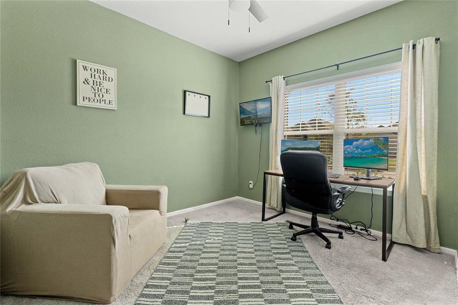 Downstairs office area featuring carpet floors, ceiling fan, and large window facing front of home