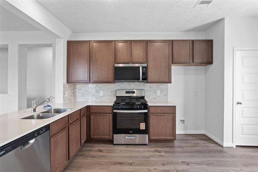 This open kitchen boasts soaring ceilings, Woodmont Cody cabinets in Toffee stain, Granite countertops in Caramel Cream, stainless steel Whirlpool appliances, a modern Daltile backsplash, recessed lighting, and a pantry—all overlooking the expansive family room.