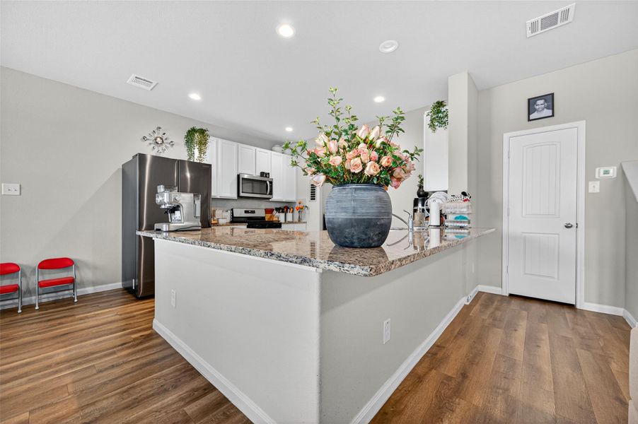 This kitchen features modern white cabinetry, stainless steel appliances, and a spacious granite countertop with a breakfast bar. The open layout and wood flooring add warmth, complemented by a large vase centerpiece.