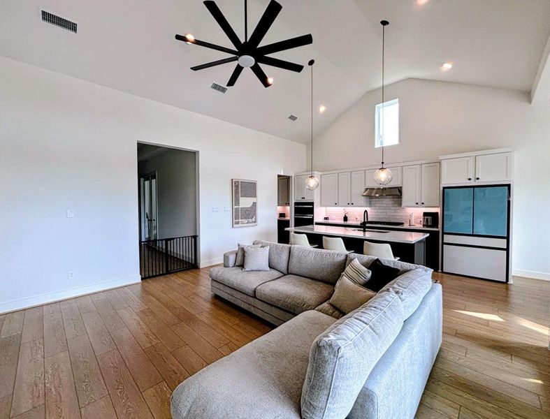 Open-concept living space with high ceilings, featuring a modern ceiling fan, and a spacious kitchen with white cabinetry and a large island. Natural light fills the room, creating a bright and inviting atmosphere.