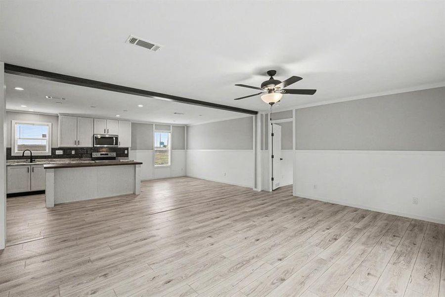 Spacious open-concept room featuring wood-look flooring, a contemporary ceiling fan with integrated lighting, and a kitchen area with light-toned cabinetry, stainless steel appliances, and dark countertops