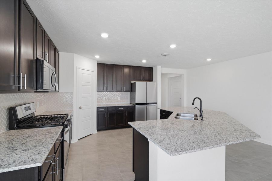 This spacious kitchen boasts soaring ceilings, Woodmont Belmont cabinets in a Espresso stain, Granite White Napoli countertops, stainless steel Whirlpool appliances, a modern Daltile backsplash, recessed lighting, an extended breakfast bar countertop, and a pantry—all overlooking the expansive family room.