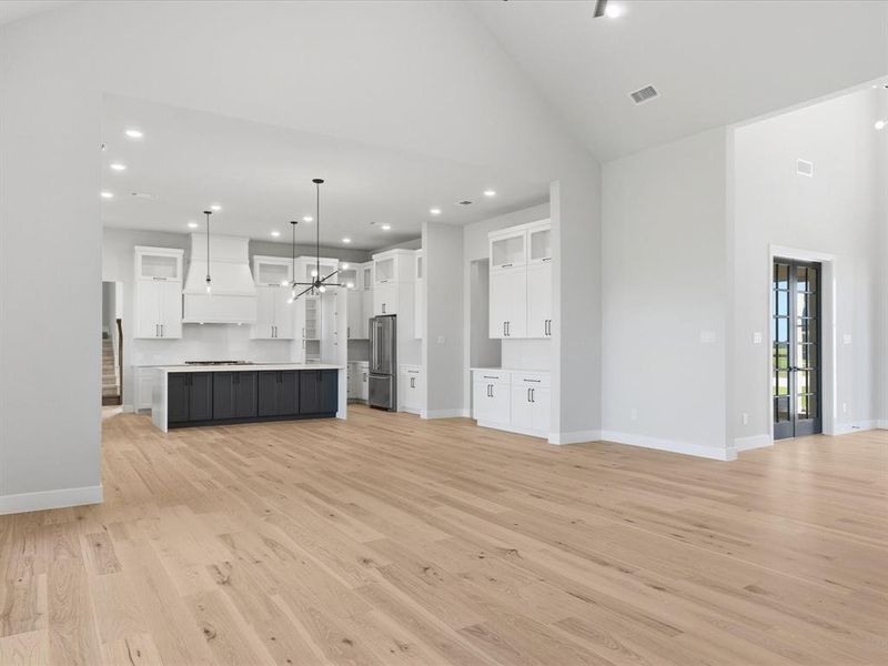 Open-concept living space featuring wood-finish flooring, a contemporary kitchen with a large island and white shaker cabinetry, an integrated stainless steel refrigerator, and vaulted ceilings