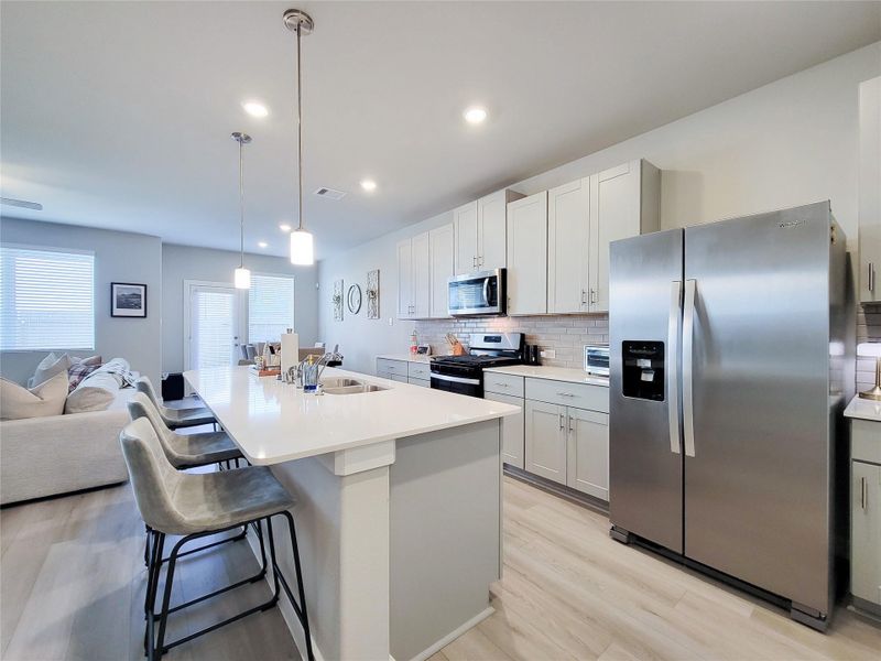 This modern kitchen features sleek stainless steel appliances, white cabinetry, and a spacious island with seating. The open layout seamlessly connects to the living area, enhanced by natural light from large windows. Ideal for entertaining and everyday living.
