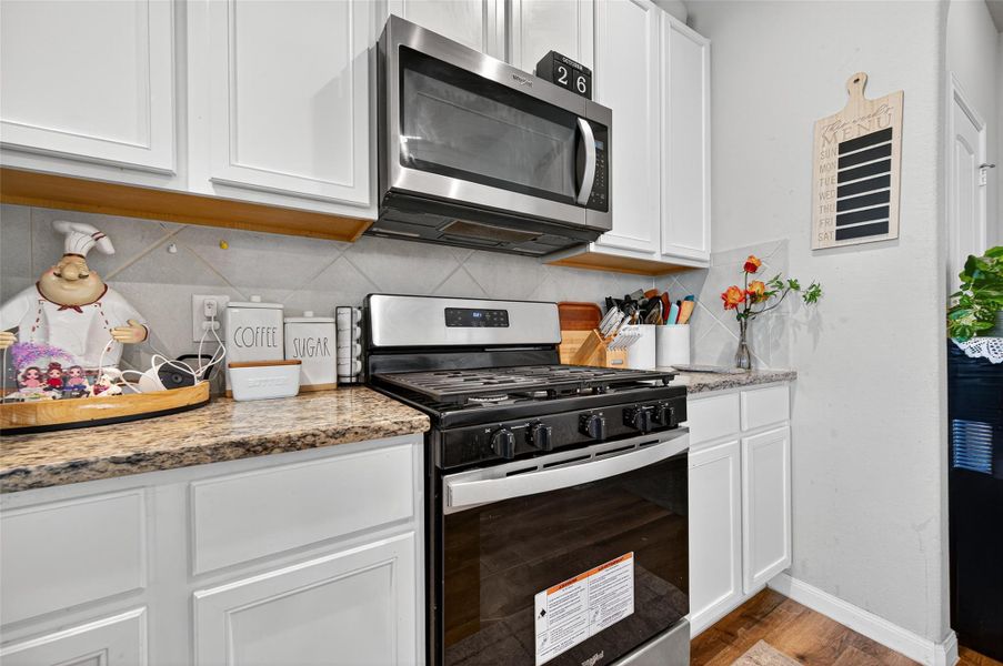 This kitchen features modern stainless steel appliances, including a built-in microwave and gas stove. It has granite countertops, white cabinetry, and a cozy layout with decorative touches.