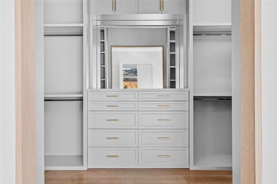 This walk-in closet boasts custom-built cabinetry with ample storage, elegantly accented by Signature Hardware brass cabinet pulls. A clerestory window allows natural light to filter in, enhancing the airy ambiance. The rich white oak hardwood floors add warmth and sophistication, creating a beautifully organized and inviting dressing space.