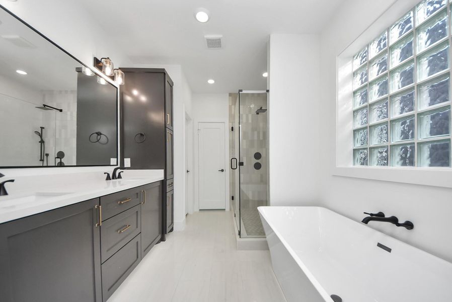 A spa-inspired bathroom with a sleek double vanity, expansive mirror, and generous storage. A glass-enclosed shower, freestanding tub, and glass block windows allow natural light to enhance the airy, modern design.