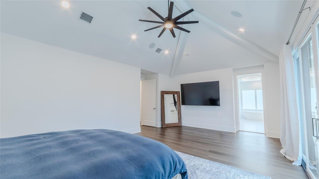 This spacious bedroom features a high vaulted ceiling with a modern fan, hardwood floors, and a large wall-mounted TV. It includes sliding glass doors for natural light and access to an outdoor area, with a glimpse of an en-suite bathroom.