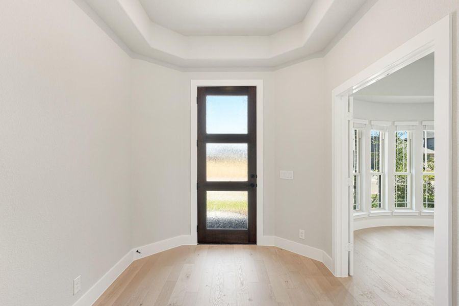 Entryway with dark wood glass-panel door, tray ceiling, and light hardwood floors leading to a bay window room. Entryway with dark wood glass-panel door, tray ceiling, and light hardwood floors leading to a bay window room.