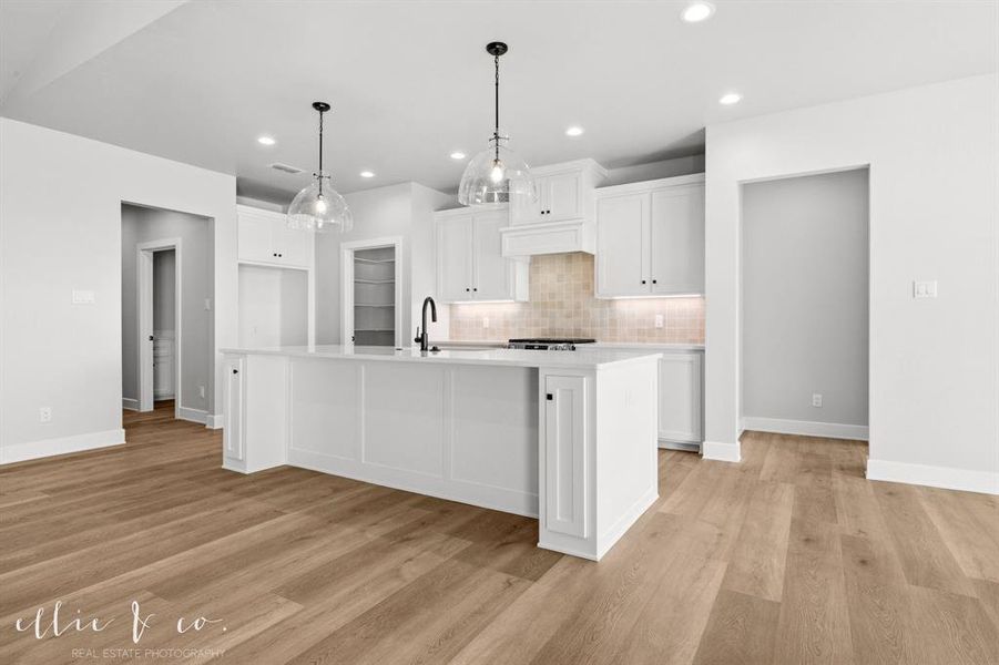 Kitchen featuring backsplash, white cabinetry, an island with sink, decorative light fixtures, and light wood-style floors