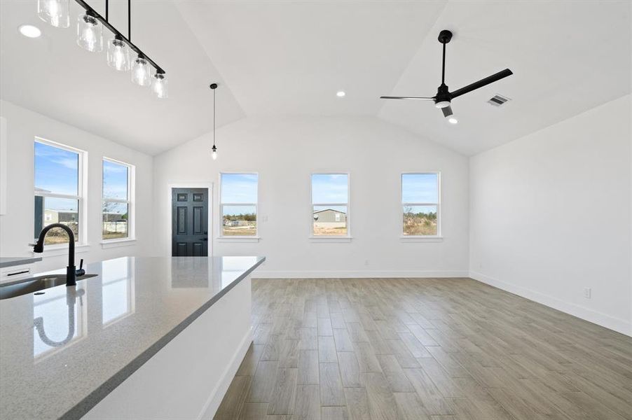 This is a bright, modern open-plan space featuring a high vaulted ceiling with recessed lighting, contemporary light fixtures, and a ceiling fan. The room has large windows providing ample natural light and views of the surrounding area. Elegant wood-look flooring extends throughout, and the kitchen area includes a sleek island with a built-in sink and a modern faucet. The neutral color palette offers a versatile backdrop for personalization.