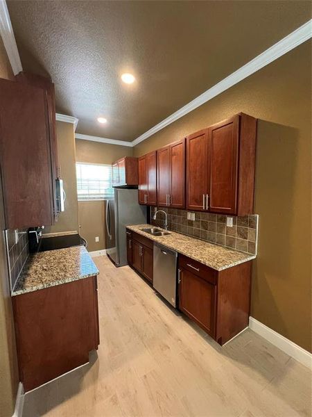 Kitchen with decorative backsplash, light stone countertops, appliances with stainless steel finishes, light wood finished floors, and a textured ceiling Kitchen with decorative backsplash, light stone countertops, appliances with stainless steel finishes, light wood finished floors, and a textured ceiling