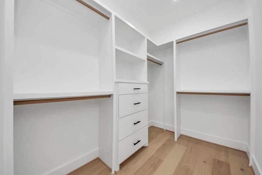 A spacious walk-in closet with custom white shelving, multiple hanging rods, and built-in drawers. Light wood flooring and bright lighting create a clean, organized space.
