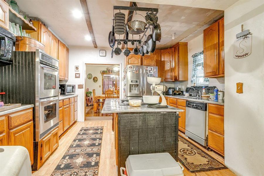 Kitchen with appliances with stainless steel finishes, light wood finished floors, a kitchen island, beam ceiling, and brown cabinetry Kitchen with appliances with stainless steel finishes, light wood finished floors, a kitchen island, beam ceiling, and brown cabinetry