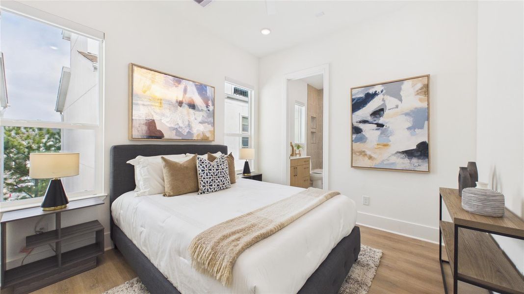 This bright bedroom features a cozy queen-sized bed with stylish cushions and a soft throw blanket. Large windows provide ample natural light, complemented by modern art pieces and sleek lighting. A doorway leads to an en-suite bathroom, enhancing convenience and privacy.