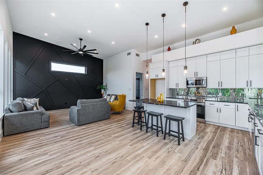 Modern, open-concept living space featuring a stylish kitchen with white cabinetry and a central island with bar seating, a cozy living area with distinctive dark accent wall and contemporary finishes, high ceilings with recessed lighting, and elegant vinyl wood flooring throughout.