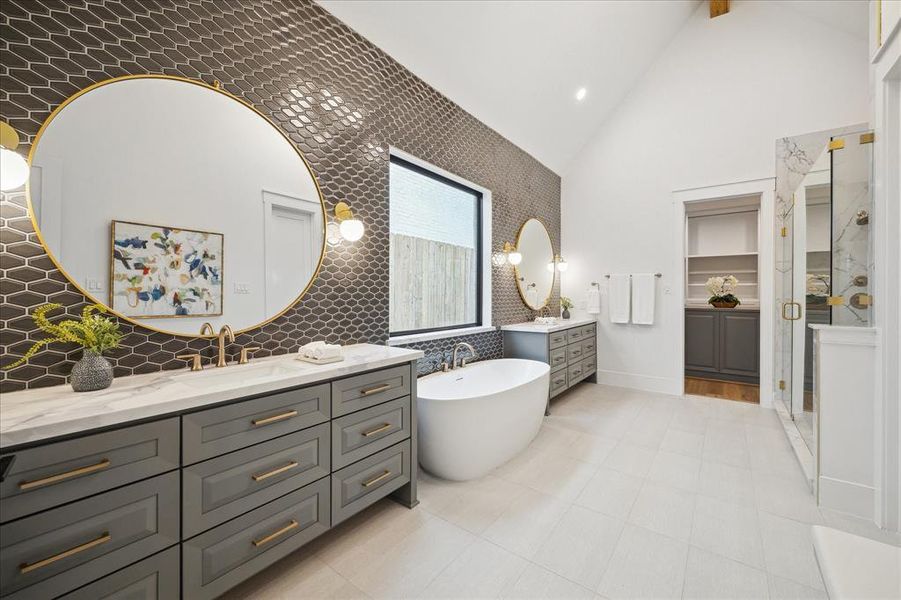 The primary bath features a vaulted ceiling, dual vanities, quartz countertops, a stand alone tub, mosaic wall tile, decorative mirrors and large textured floor tiles.